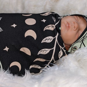 May include: A baby swaddled in a black and green swaddle with a moon and leaf pattern. The baby is wearing a matching hat with a moth design.