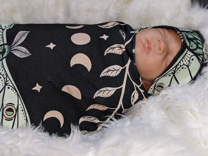 May include: A baby swaddled in a black and green swaddle with a moon and leaf pattern. The baby is wearing a matching hat with a moth design.