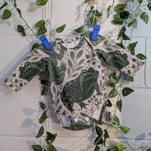 May include: A long-sleeved baby onesie with a light pink background and a green frog print. The onesie has a frog with a detailed pattern on the front and smaller frogs on the sleeves.