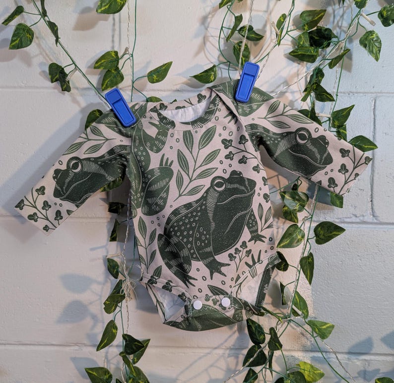 May include: A long-sleeved baby onesie with a light pink background and a green frog print. The onesie has a frog with a detailed pattern on the front and smaller frogs on the sleeves.