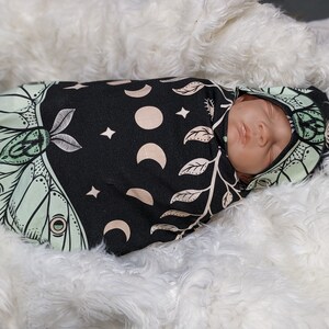 May include: A baby swaddled in a black and green swaddle with a moth and moon design. The swaddle has a hood and is made of a soft, stretchy fabric.