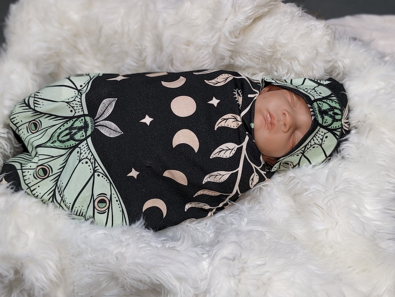 May include: A baby swaddled in a black and green swaddle with a moth and moon design. The swaddle has a hood and is made of a soft, stretchy fabric.