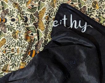 May include: A blanket with a cream-colored background featuring a pattern of mushrooms, bees, and foliage in shades of green, yellow, and brown. The blanket has a black satin-like underside with the word "eithy" in white script.