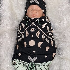 May include: A black swaddle with a green moth, moon phases, stars, and an eye design. A baby is wearing the swaddle and a matching black and green hat.
