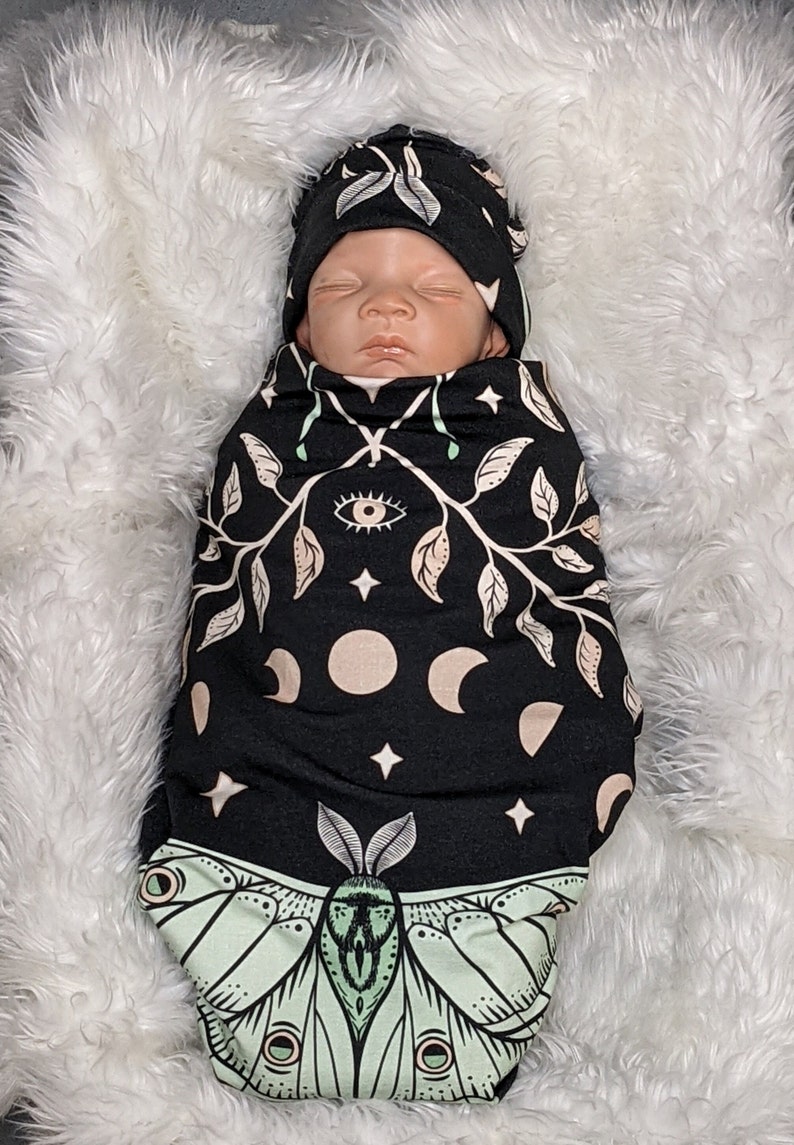 May include: A black swaddle with a green moth, moon phases, stars, and an eye design. A baby is wearing the swaddle and a matching black and green hat.