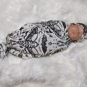 May include: A baby swaddled in a white and black swaddle with a moth and floral pattern. The baby is wearing a matching black and white hat.