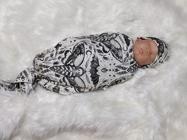 May include: A baby swaddled in a white and black swaddle with a moth and floral pattern. The baby is wearing a matching black and white hat.