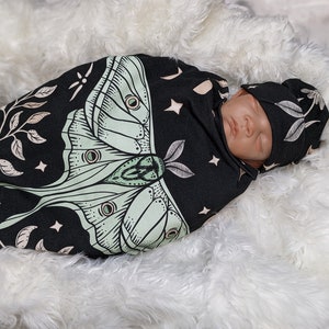 May include: A black and cream swaddle with a green moth design and stars. A baby is sleeping inside the swaddle wearing a matching hat.