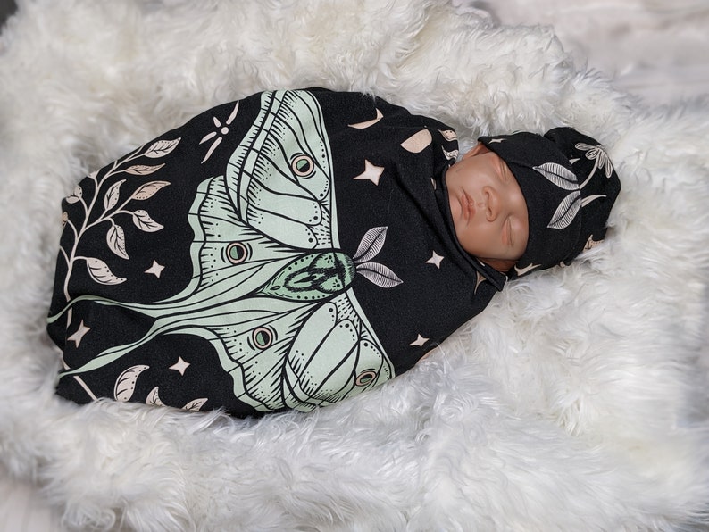 May include: A black and cream swaddle with a green moth design and stars. A baby is sleeping inside the swaddle wearing a matching hat.
