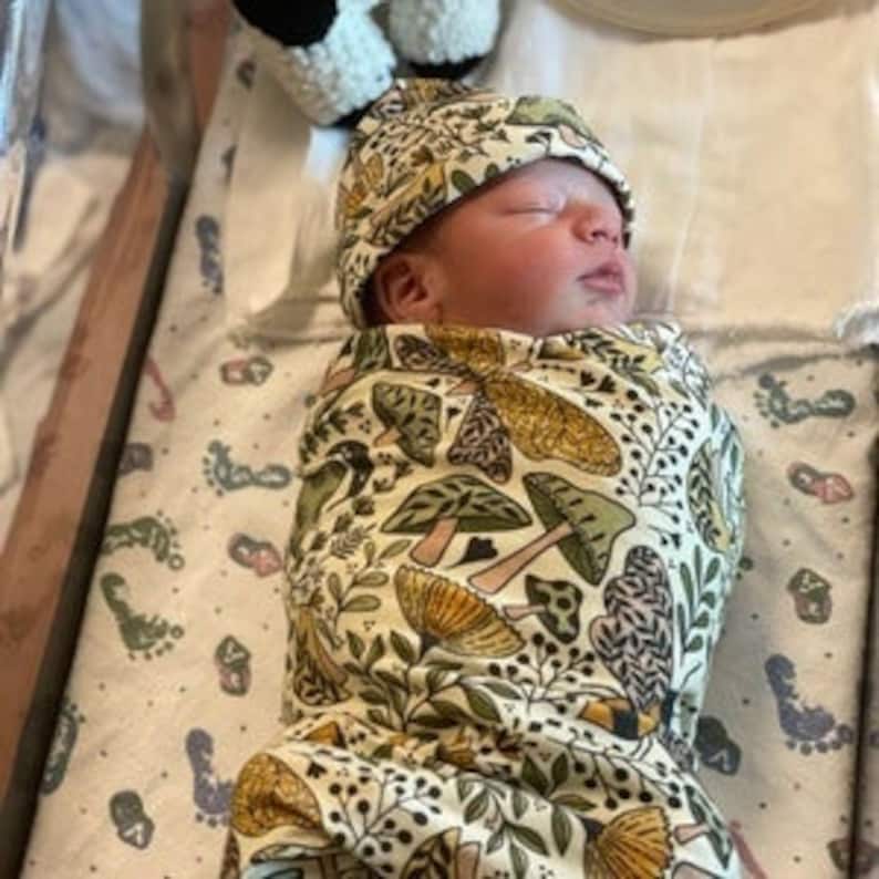 May include: A newborn baby swaddled in a white and green swaddle with a mushroom and leaf pattern. The baby is wearing a matching mushroom and leaf patterned hat.