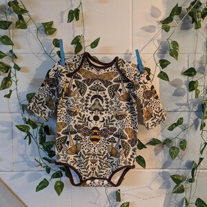 May include: A long-sleeved baby onesie with a beige background and a colourful pattern of mushrooms, bees, and flowers.