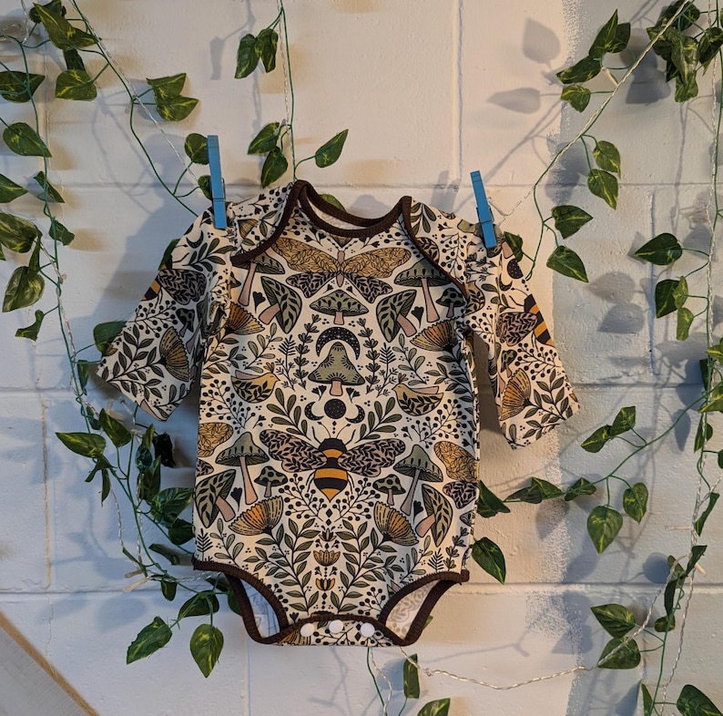 May include: A long-sleeved baby onesie with a beige background and a colourful pattern of mushrooms, bees, and flowers.