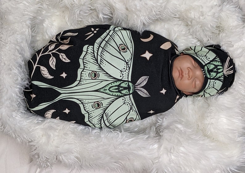 May include: A black and green swaddle with a moth design and a matching hat. The swaddle has a white crescent moon and stars on it. The hat has a moth design on it.