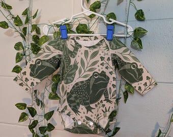 May include: A long-sleeved baby bodysuit with a beige background and a repeating pattern of green frogs and foliage. The bodysuit is hanging on a white hanger with blue clips, and is surrounded by artificial greenery.