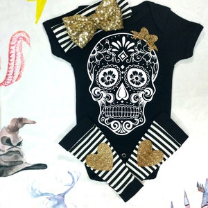 May include: Black and white baby onesie with a sugar skull design. The onesie has black and white striped leg warmers with gold glitter hearts. A gold glitter bow is included.
