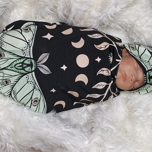 May include: A black and green swaddle with a moth and moon design. A baby is sleeping in the swaddle with a matching hat.