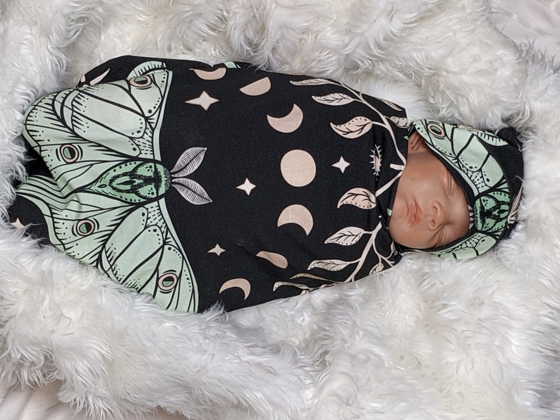 May include: A black and green swaddle with a moth and moon design. A baby is sleeping in the swaddle with a matching hat.