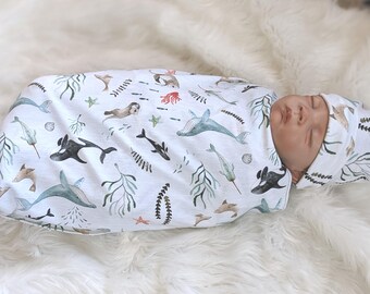May include: A baby swaddled in a white blanket and matching cap, both adorned with an aquatic-themed pattern. The design features whales, sea creatures, and seaweed in shades of blue, gray, and brown. The baby is lying on a fluffy white surface.