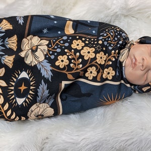 May include: A baby swaddled in a black and blue swaddle with a floral and celestial pattern. The swaddle features a white eye with a star in the centre, a crescent moon, and a sun. The baby is wearing a matching hat.