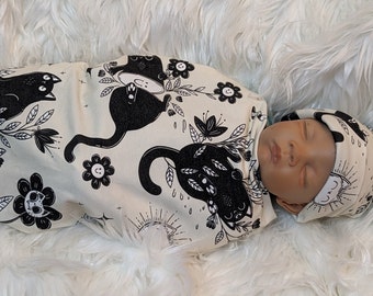 May include: A newborn baby is swaddled in a white and black swaddle with a black cat and flower pattern. The baby is wearing a matching black and white hat with a sun and moon design.