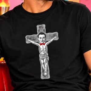 May include: A black short-sleeved t-shirt featuring a white graphic print of a person with a red bow tie depicted on a cross. The person is shown with arms outstretched, wearing a loincloth, and the design includes intricate line work details.