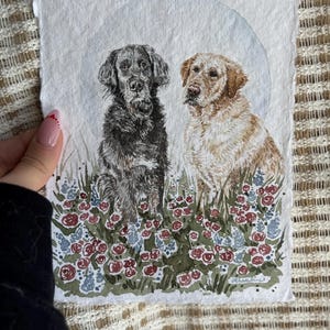 以下が含まれることがあります： 2匹の犬の水彩画。1匹は黒、もう1匹は金色で、花畑に座っています。このアートワークは、淡い青色の背景と赤、青、緑の花の要素を備えた、柔らかく絵画的なスタイルです。