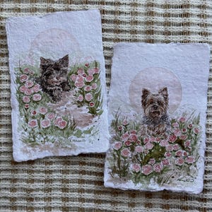 以下が含まれることがあります： 庭にいるヨークシャーテリアの2つの水彩画。各絵画は、ピンクのバラと緑の葉に囲まれた犬を特徴とし、柔らかい円形の背景があります。アートワークはテクスチャード加工された紙に描かれています。