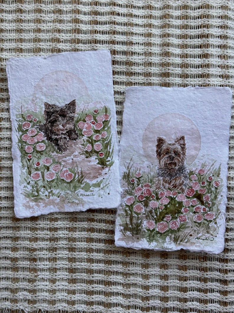 以下が含まれることがあります： 庭にいるヨークシャーテリアの2つの水彩画。各絵画は、ピンクのバラと緑の葉に囲まれた犬を特徴とし、柔らかい円形の背景があります。アートワークはテクスチャード加工された紙に描かれています。