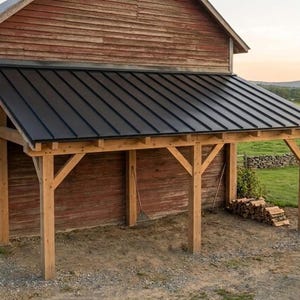 May include: A wooden lean-to structure attached to a red barn, featuring a black metal roof with vertical seams. The open-sided shelter is supported by sturdy timber posts and beams, providing a covered area for storage and outdoor equipment.