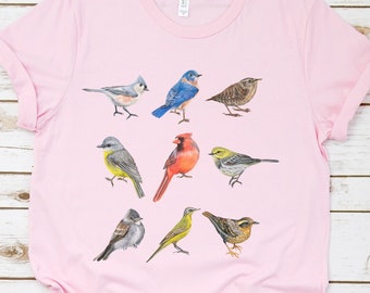 May include: Pink t-shirt with a colorful design of various birds. The birds are illustrated in a realistic style and include a bluebird, cardinal, chickadee, and more.