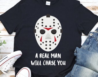May include: A dark blue t-shirt with a white hockey mask graphic and the text "A real man will chase you". The shirt is laid out on a wooden surface with a pair of white sneakers and blue jeans.