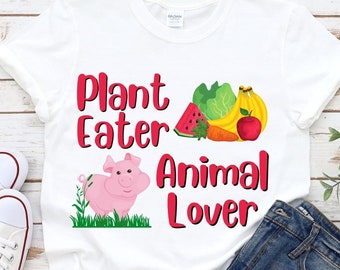 May include: White t-shirt with the text "Plant Eater Animal Lover" in pink. The shirt features a cartoon pig and a variety of fruits and vegetables.