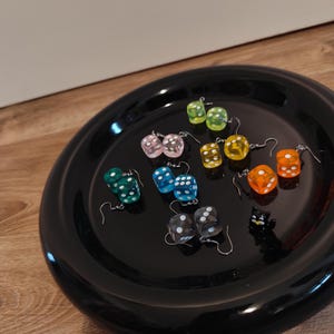 Könnte beinhalten: Eine Sammlung von Würfelohrringen in verschiedenen Farben, darunter Grün, Rosa, Blau, Gelb, Orange und Schwarz. Die Ohrringe werden auf einem schwarzen Teller präsentiert, der ihre vielfältigen Designs und Farben zeigt. Die Würfel haben weiße Punkte.