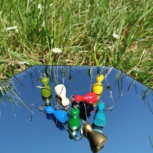 以下が含まれることがあります： 7つのカラフルな木製ゲームピースのイヤリングのセット。イヤリングは人の形をしていて、緑、青、赤、黄、金など様々な色で塗られています。イヤリングは鏡面上に飾られています。