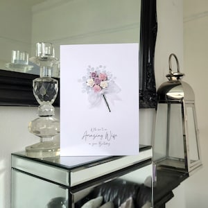 Peut inclure: Une carte blanche avec un bouquet de fleurs roses et blanches et le texte "A little love to an Amazing Wife on your Birthday".