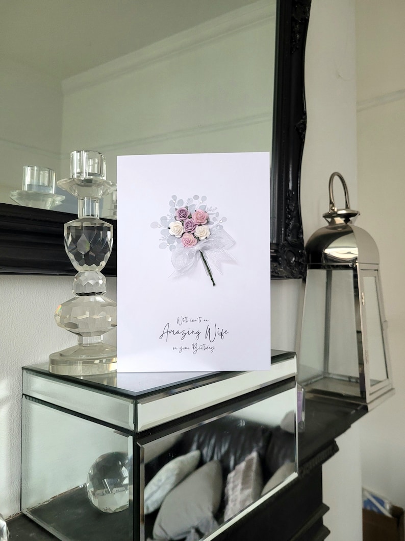 Peut inclure: Une carte blanche avec un bouquet de fleurs roses et blanches et le texte "A little love to an Amazing Wife on your Birthday".