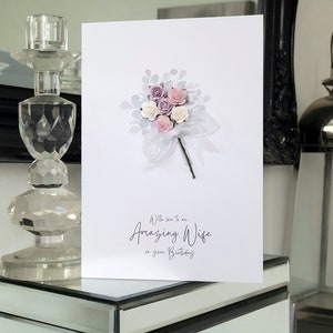 Peut inclure: Une carte blanche avec un bouquet de fleurs roses et blanches avec des accents argent&eacute;s. La carte dit "Avec amour pour une femme extraordinaire pour ton anniversaire."