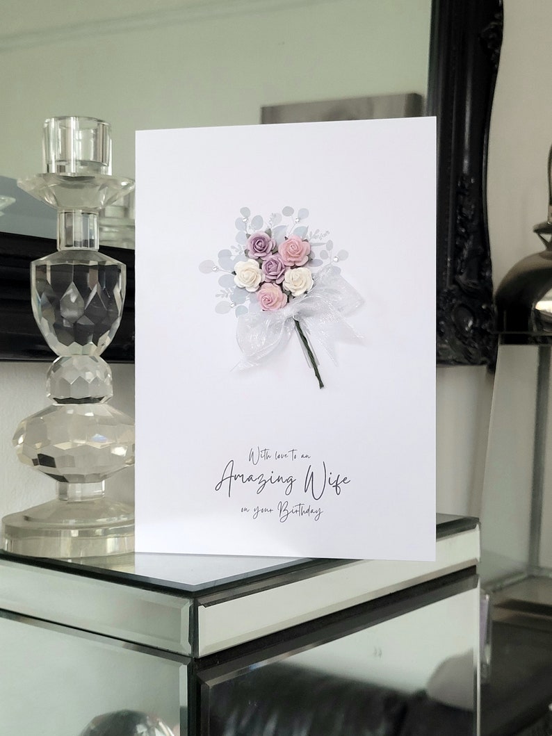 Peut inclure: Une carte blanche avec un bouquet de fleurs roses et blanches avec des accents argent&eacute;s. La carte dit "Avec amour pour une femme extraordinaire pour ton anniversaire."