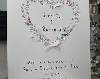 May include: A white wedding card with a heart-shaped floral design, adorned with butterflies and the names "Archie & Rebecca." The card includes the text "With love to a wonderful Son & Daughter-in-Law on your Wedding Day."