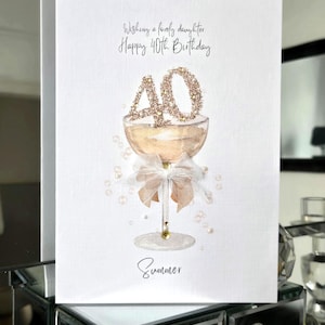 May include: A white birthday card with a champagne glass design. The card reads "Wishing a lovely daughter Happy 40th Birthday" with the name "Summer" at the bottom. The number "40" is decorated with gold glitter.