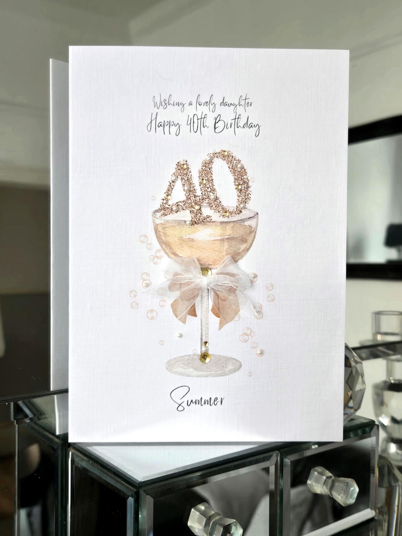 May include: A white birthday card with a champagne glass design. The card reads "Wishing a lovely daughter Happy 40th Birthday" with the name "Summer" at the bottom. The number "40" is decorated with gold glitter.