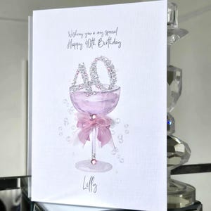 May include: A white greeting card with the text "Wishing you a very special Happy 40th Birthday." The card features a pink champagne glass with a "40" made of silver glitter. A pink bow and the name "Lilly" are also on the card.