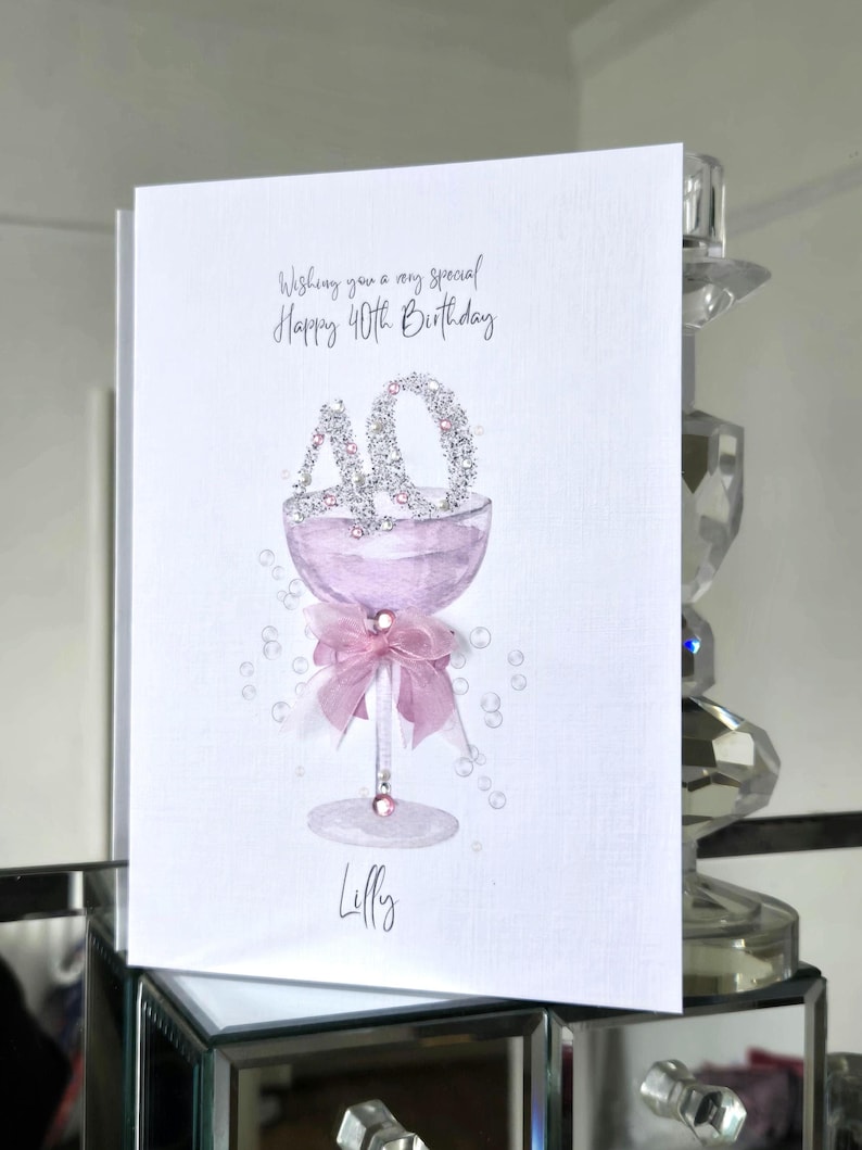 May include: A white greeting card with the text "Wishing you a very special Happy 40th Birthday." The card features a pink champagne glass with a "40" made of silver glitter. A pink bow and the name "Lilly" are also on the card.