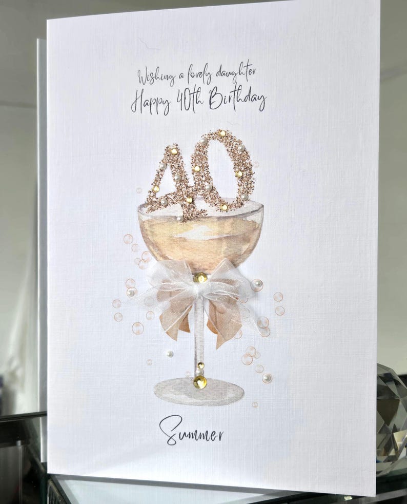 May include: A birthday card with a champagne glass design. The card reads "Wishing a lovely daughter Happy 40th Birthday" and includes the name "Summer". The number "40" is decorated with glitter.