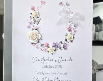 May include: A white wedding card with a floral wreath design. The wreath is composed of white, pink, and purple flowers, with butterflies and a white ribbon. The card reads "Christopher & Amanda" and "25th July 2025".