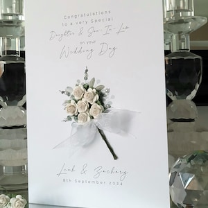 Puede incluir: Tarjeta de boda blanca con un ramo de flores blancas y una cinta blanca. La tarjeta dice "Felicidades a una hija y un yerno muy especiales por su d&iacute;a de boda" y "Leah & Zachary 8 de septiembre de 2024".