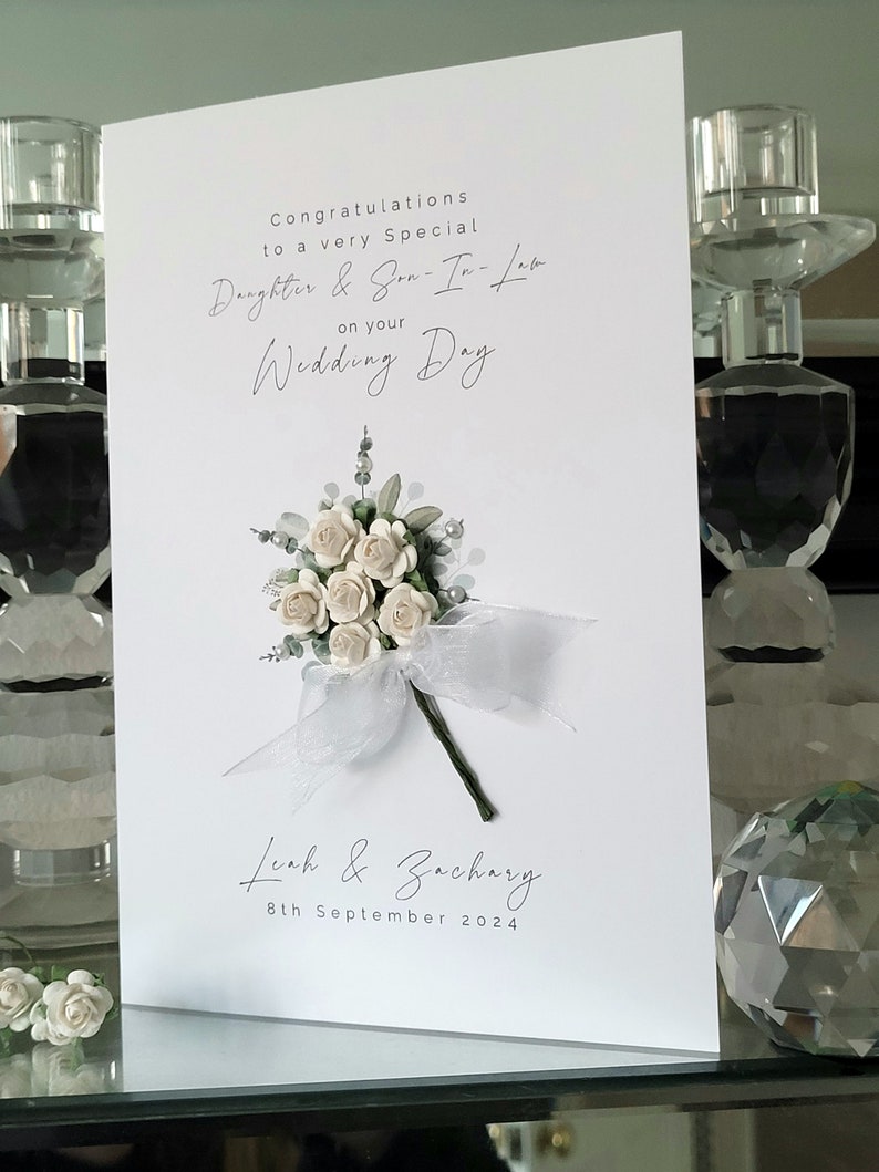 Puede incluir: Tarjeta de boda blanca con un ramo de flores blancas y una cinta blanca. La tarjeta dice "Felicidades a una hija y un yerno muy especiales por su d&iacute;a de boda" y "Leah & Zachary 8 de septiembre de 2024".