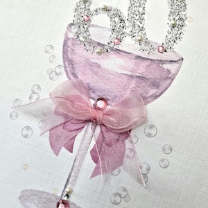 May include: A greeting card featuring a watercolor-style champagne glass in shades of pink. The glass is adorned with a sparkly "60" and a pink bow. Embellished with rhinestones and faux pearls, the card has a celebratory theme.
