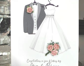 May include: A white card with a gray suit and a white dress on hangers. The suit has a pink flower on the lapel. The dress has a pink flower bouquet. The text reads "Congratulations on your Wedding day Adam & Rebecca 1st October 2022".