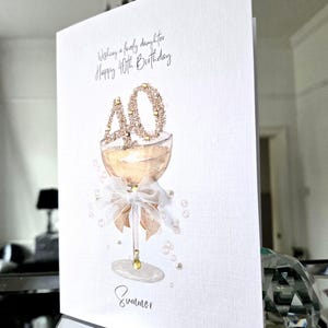 May include: A white birthday card with the text "Wishing a lovely daughter Happy 40th Birthday." The card features a champagne glass with a "40" made of gold glitter. A white bow and pearls decorate the glass. The name "Summer" is written at the bottom.